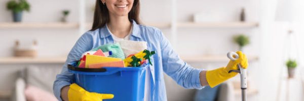 Portrait of smiling millennial lady holding bucket with cleaning supplies and mop, posing and looking at camera standing in living room. Professional cleaning service specialist wearing rubber gloves