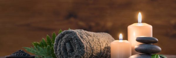 Brown towel on green fern with candles and black hot stone on wooden background. Hot stone massage setting lit by candles. Hot stone therapy for one person with candle light. Beauty spa treatment and relax concept.